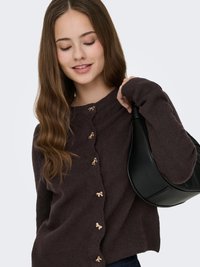 Junge Frau mit langen braunen Haaren, die eine dunkelbraune Strickjacke mit goldenen Schleifenknöpfen trägt und eine schwarze Lederhandtasche über der Schulter hält.