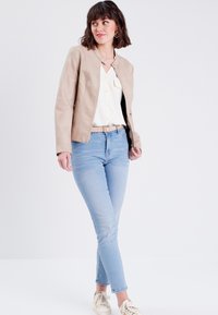 Veste en cuir beige avec un col rond, une fermeture éclair et des détails de coutures. Associée à une chemise blanche et un jean skinny bleu clair.