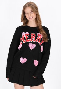 Pulover negru cu "MERRY" scris cu litere roșii, decorat cu inimioare din sequins roz. Materialul pare moale și texturat. Este asortat cu o fustă neagră plisată.
