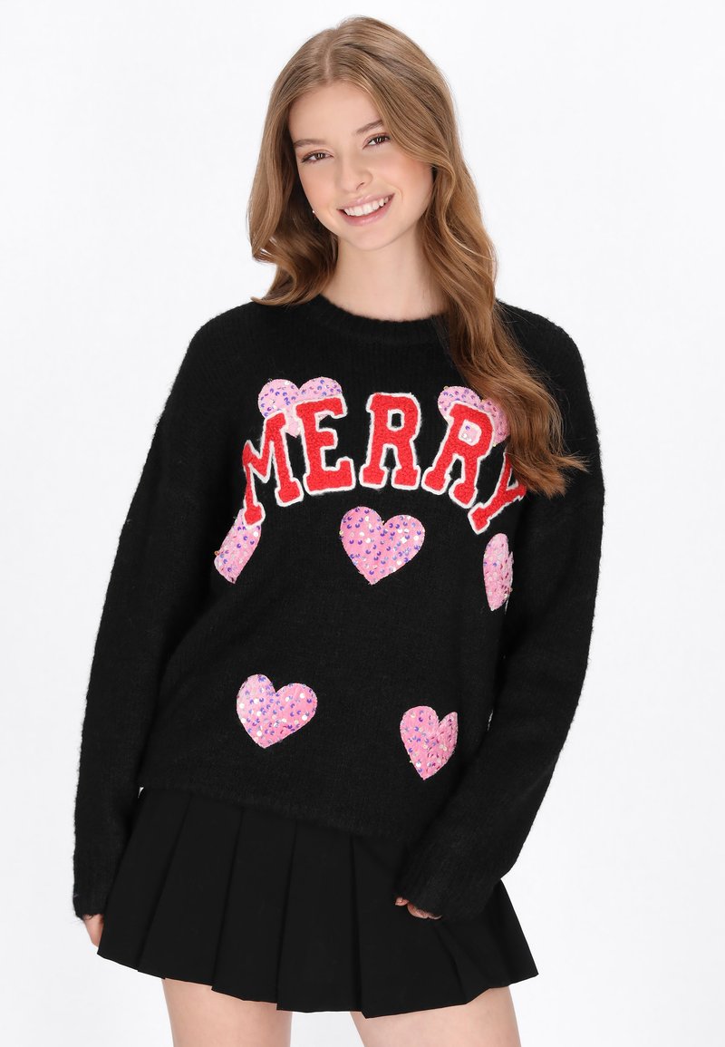 Pulover negru cu "MERRY" scris cu litere roșii, decorat cu inimioare din sequins roz. Materialul pare moale și texturat. Este asortat cu o fustă neagră plisată.