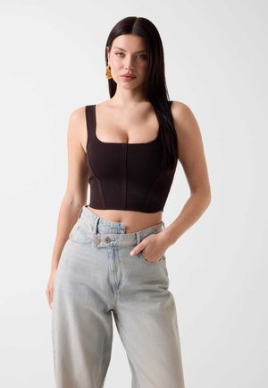 Femme aux longs cheveux foncés, portant des boucles d'oreilles créoles dorées, un débardeur court noir et un jean bleu clair taille haute avec un design inhabituel à double bouton.