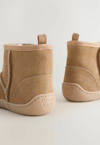 Helle Wildleder-Stiefeletten mit weicher Fütterung, strukturierten Oberflächen und Zuglaschen. Merkmale ein Bärenemblem auf der beigen Gummisohle für zusätzlichen Detail.
