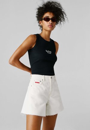 Kobieta z kręconymi włosami, ubrana w czarną, bezrękawnikową bluzkę z logo Red Bull Racing, białe szorty z logo RBR oraz czarne okulary przeciwsłoneczne, stojąca na szarym tle.