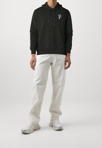 Felpa nera con tasca frontale e grafica bianca, abbinata a jeans bianchi larghi e sneakers argentate. Design semplice e casual.