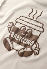 Fehér póló, amelyen egy barna rajzfilmszerű kávéscsésze grafika látható. A csésze mosolygó arccal, karokkal rendelkezik, és a "BABYCCINO" szó hangsúlyosan van nyomtatva.