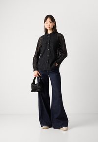 Black lace shirt with button closure, two chest pockets; paired with wide-leg dark denim jeans and a small black handbag.
