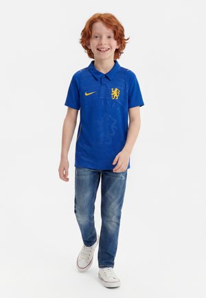 Garçon roux souriant marchant portant un maillot bleu du club de football Chelsea, un jean bleu et des baskets blanches sur fond blanc.