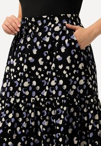 Musta hame, jossa on valkoisia ja laventelinvärisiä abstrakteja täpliä, joustava vyötärö naruilla ja sivutaskut. Sisältää kerroksellisen röyhelömuotoilun.