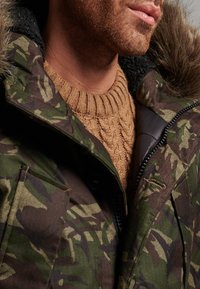 Parka de camuflaje con capucha forrada de piel sintética, con cierre de cremallera y bolsillos con cremallera, llevada sobre un suéter de punto texturizado color beige.