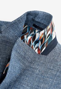 Col blanc en tissu texturé bleu avec une doublure intérieure à motifs multicolores présentant des formes ondulées dans des tons marron, bleu et blanc.