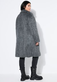 Abrigo largo de piel gris con cuello, que presenta un exterior texturizado. Combinado con pantalones negros ajustados y botas negras gruesas.