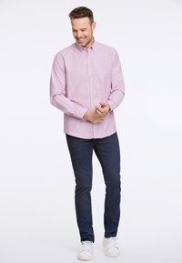 Camisa de botones de color rosa claro con un bolsillo, usada sobre pantalones vaqueros azul oscuro y zapatillas blancas, con mangas largas y dobladillo curvado.