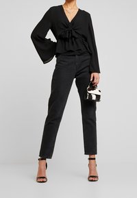 Černá halenka s dlouhým rukávem, výstřihem do V a uzlovým detailem v pase, doplněná černými džíny s vysokým pasem a malou černo-bílou kabelkou.