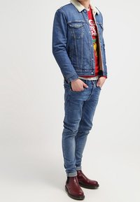 Veste en denim avec col en fausse fourrure, sweat-shirt rouge avec imprimé, jean bleu et bottines bordeaux à semelle noire. Coupe slim, design décontracté.
