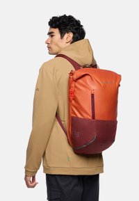 Mochila naranja y burdeos con un diseño de rollo, hecha de tela texturizada, que presenta un bolsillo con cremallera y correas ajustables.
