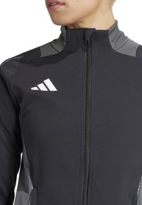 Veste de sport noire avec des accents gris, col montant, fermeture éclair intégrale et logo blanc sur la poitrine. Fabriquée en matériau lisse et extensible.