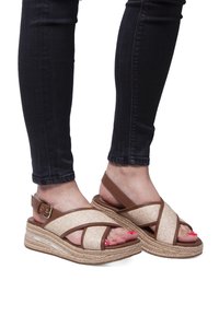 Sandalias espadrilles beige con tiras cruzadas, detalles marrones y suelas de yute trenzado; exhibidas en pies con pantalones oscuros y uñas de los pies pintadas.
