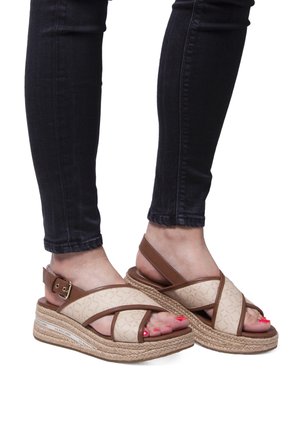 Sandales espadrilles beige avec des brides croisées, des accents marron et des semelles en jute tressé ; portées avec un jean foncé et des ongles de pieds vernis.
