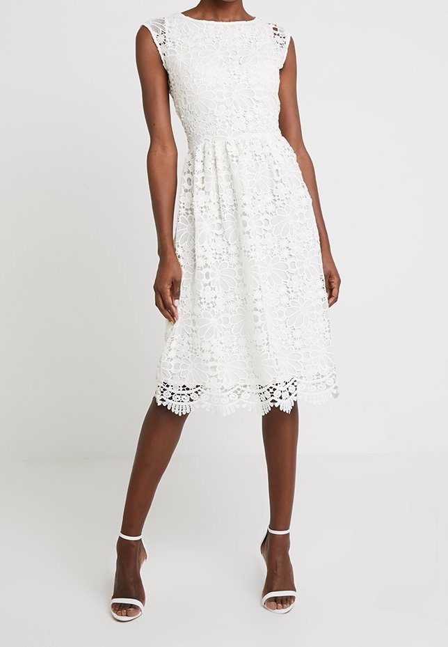 Robe en dentelle blanche avec col rond, sans manches, motifs floraux et ourlet festonné. Associée à des sandales à talons blanches.