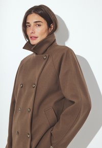 Woman wearing a brown double-breasted coat with raised collar standing against a white wall, hands in pockets.