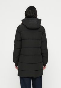 Sort vattert jakke med hette, horisontale sømmer og en litt forlenget fasong. Mykt, teksturert materiale med elastiske mansjetter.