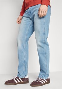 Hellblaue Denim-Jeans mit geradem Schnitt, sichtbaren Nähten und einem ausgewaschenen Finish, kombiniert mit dunkelroten Sneakern mit weißen Akzenten.