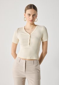 Haut à manches courtes côtelé de couleur crème clair avec un col en V et des boutons, associé à un pantalon taillé beige clair. Texture lisse.