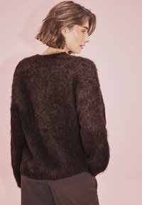Rudas, pūkuotas mohair megztinis laisvu siluetu, plačiomis rankogaliais ir apvalia iškirpte, dėvimas ant tamsių kelnių prieš rožinį foną.