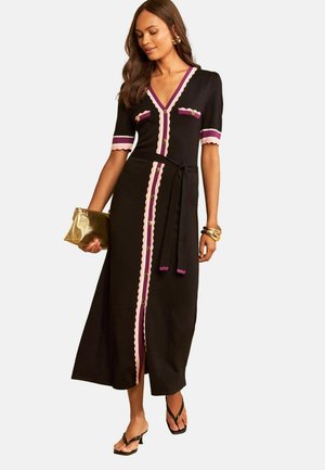 Kvinde iført en sort midi-kjole med pink og lilla kant, knapper foran, bælte, holder en gylden clutch, iført sorte sandaler og gyldne armbånd.