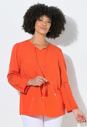 Kvinde med krøllet hår iført en orange bluse med lange ærmer med frynsede bindebånd og hvide bukser, stående mod en hvid baggrund.