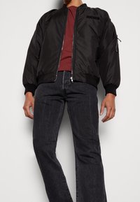 Chaqueta bomber negra con puños y cuello acanalados, con cremallera frontal y bolsillo lateral. Llevada sobre una camisa burdeos y jeans oscuros.