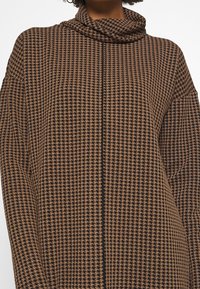 Personne portant un pull à motif pied-de-poule marron et noir avec un col roulé ample sur un fond uni.
