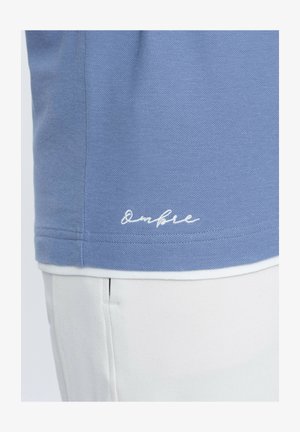 Hemd aus hellblauem Polo in atmungsaktiver Textur mit einem weißen gestickten Markennamen am Saum und kontrastierendem weißen Besatz am unteren Rand.