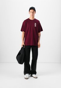 Burgundy cotton t-shirt with short sleeves and white graphic print, paired with wide black denim pants and white sneakers. Black bag in hand.