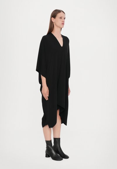 Svart oversized klänning med V-ringning och batwing-ärmar, tillverkad av mjukt tyg, med en lös passform och sidofickor, parat med svarta anklarstövlar.