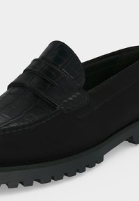 Μαύρο loafer με επάνω μέρος σε σχέδιο κροκόδειλου και ραμμένες λεπτομέρειες σε παχιά ελαστική σόλα, σε μονόχρωμο φόντο.
