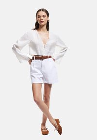 Blouse blanche courte avec des manches larges, associée à un short blanc taille haute, une ceinture marron avec boucle en argent, et des chaussures plates marron.