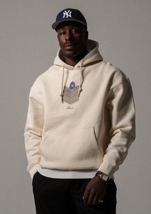 Cremefarbener Hoodie mit einer Fronttasche, Zugbändern und einem geometrischen Grafikdesign auf der Brust. Abgeschlossen mit gerippten Bündchen und Saum.