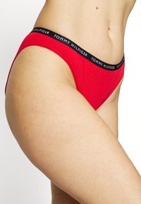 Red thong underwear with a black elastic waistband featuring "TOMMY HILFIGER" logo. Smooth fabric texture, minimalist design, and low-rise cut.