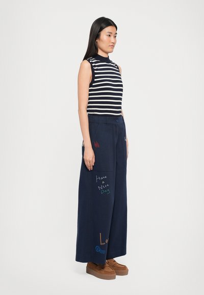Pantaloni largi de marină cu text brodat multicolor; asortați cu un top ajustat fără mâneci, cu dungi alb-negru. Pantofi cu platformă maro.