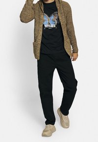 Homme portant un cardigan marron texturé, un t-shirt noir avec un imprimé papillon, un pantalon noir et des baskets beiges, posant avec la main près de la tête.