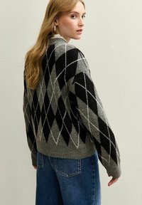 Joven mujer con cabello rubio largo, vistiendo un suéter gris y negro de rombos y jeans azules, mirando por encima de su hombro contra un fondo liso.