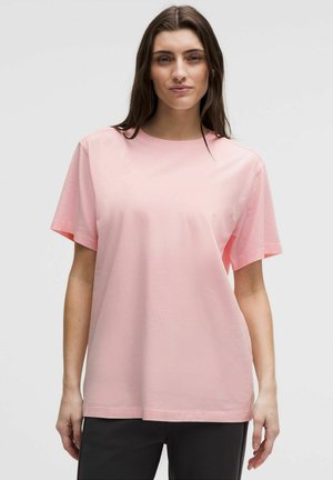 Kobieta z długimi brązowymi włosami, ubrana w jednolity jasnoróżowy t-shirt z krótkim rękawem i czarne spodnie na jednolitym tle.