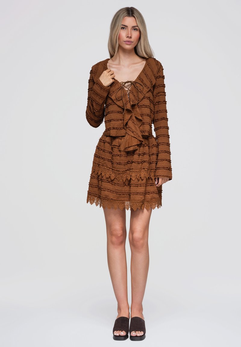 Femme portant une robe en maille marron texturée avec un devant à volants et un ourlet en dentelle, debout devant un fond clair uni avec des sandales noires à bout ouvert.