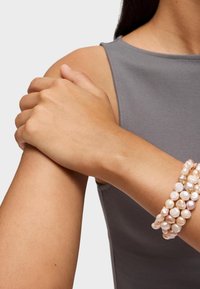 Elastyczny, wielowarstwowy bracelet z okrągłymi, iryzującymi perłami w różowych i kremowych odcieniach. Noszony na nadgarstku, w połączeniu z szarym bezrękawnikiem.