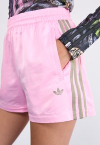 Rosa Sportshorts mit elastischem Bund, Seitenfächern und drei vertikalen Olivgrünen Streifen. Mit kleinem grauen Logo am Saum.