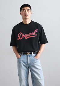 Camiseta de algodão preto com o audacioso logo vermelho "Dsquared²", combinada com jeans de denim azul claro. O modelo tem as mãos nos bolsos, em pé contra um fundo plain.