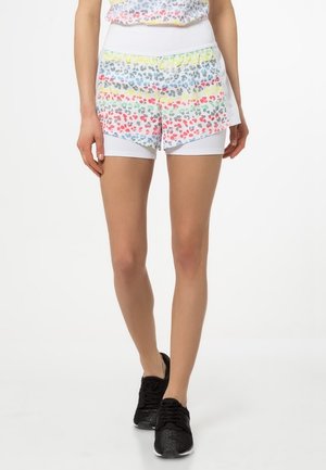 Vrouw draagt witte sportshorts met kleurrijke luipaardprint en zwarte hardloopschoenen, staand tegen een witte achtergrond.