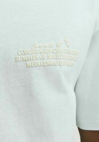  Lichtblauw katoenen T-shirt met tekstuur geborduurde tekst in crème, met details "Kopenhagen," "Zomer van 1990," en "Mediterranee."