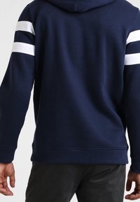 Sudadera color azul marino con rayas blancas en la parte superior de las mangas, fabricada en una tela suave. Presenta un bolsillo canguro y puños y dobladillo de canalé.